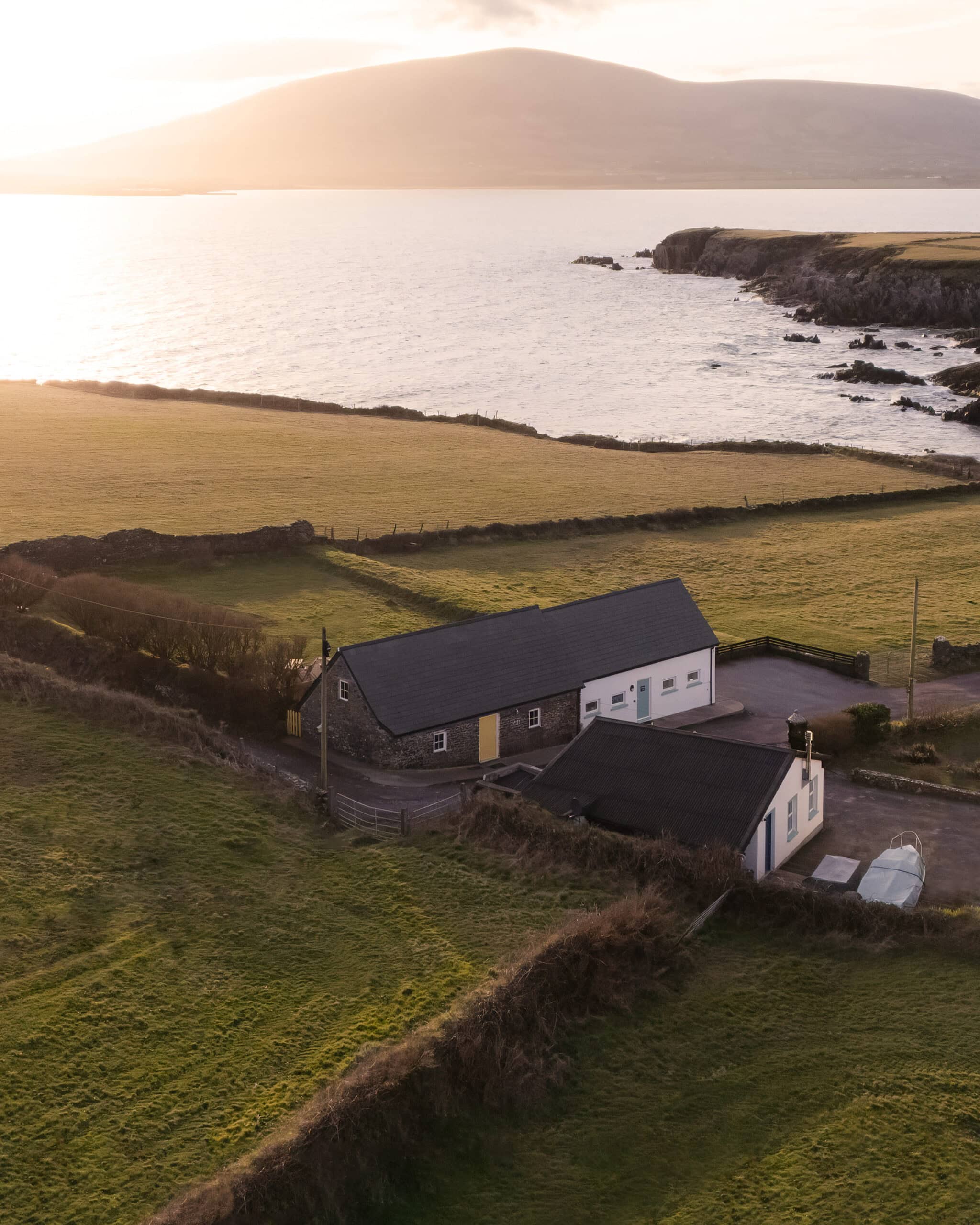 sanctuary-cottages-pipers-cottage-exterior-01 Aerial view of Pipers Cottage in Ballymore near Ventry Bay, Dingle, surrounded by green fields and overlooking the Atlantic Ocean at sunset.