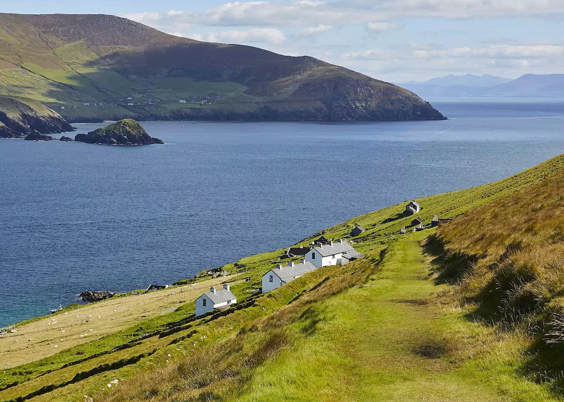 anoramic view of abandoned cottages on Great Blasket Island with sloping green hillsides, blue waters, and distant mountains of the Dingle Peninsula in the background.