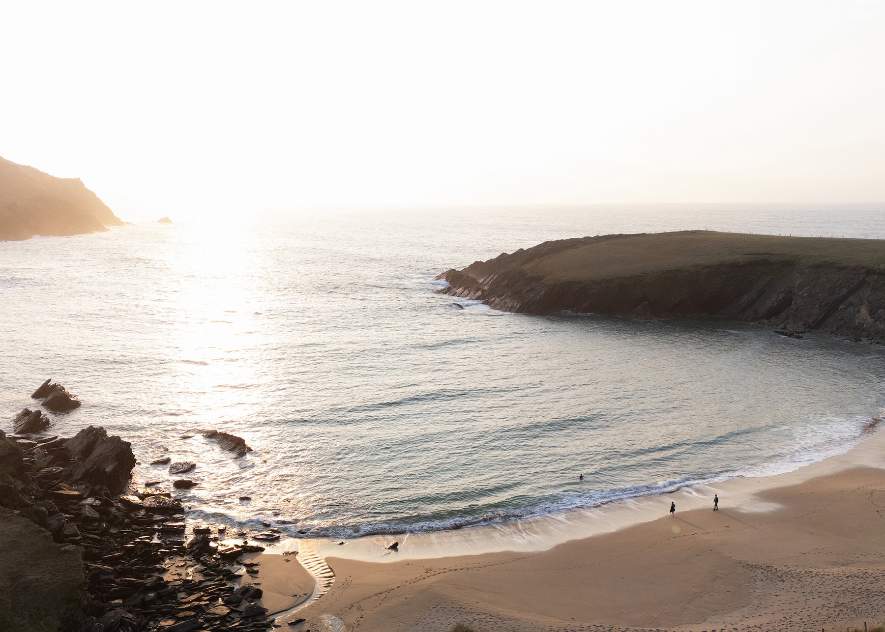 Sunset view of Clogher Strand beach on the Dingle Peninsula. Golden evening light illuminates the sheltered sandy cove surrounded by rocky headlands. A few people walk along the shoreline, with footprints visible in the sand. The beach is nestled between dramatic coastal cliffs with waves gently breaking on shore.