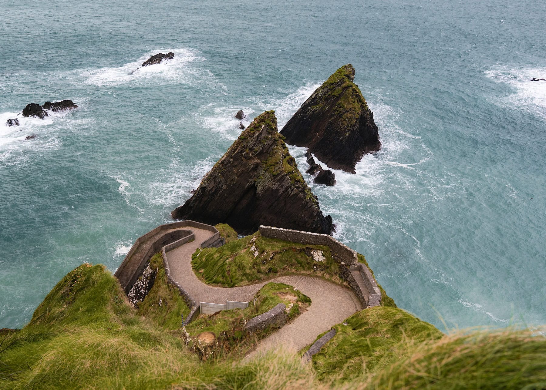 Iconic zigzagging pathway of Dunquin Pier descending from grassy cliffs to the turquoise waters below. Two distinctive triangular rock formations known as sea stacks rise from the ocean, with waves breaking around them. The winding stone-walled path creates a striking pattern against the coastal landscape of Ireland's Dingle Peninsula.