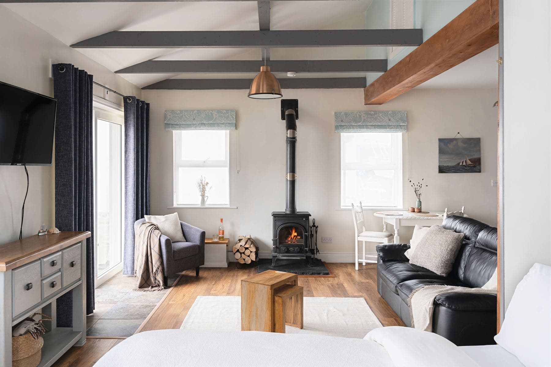 Bright open-plan living area with exposed ceiling beams, wood-burning stove, armchair, black leather sofa, and dining table by the window in The Studio at Sanctuary Cottages.