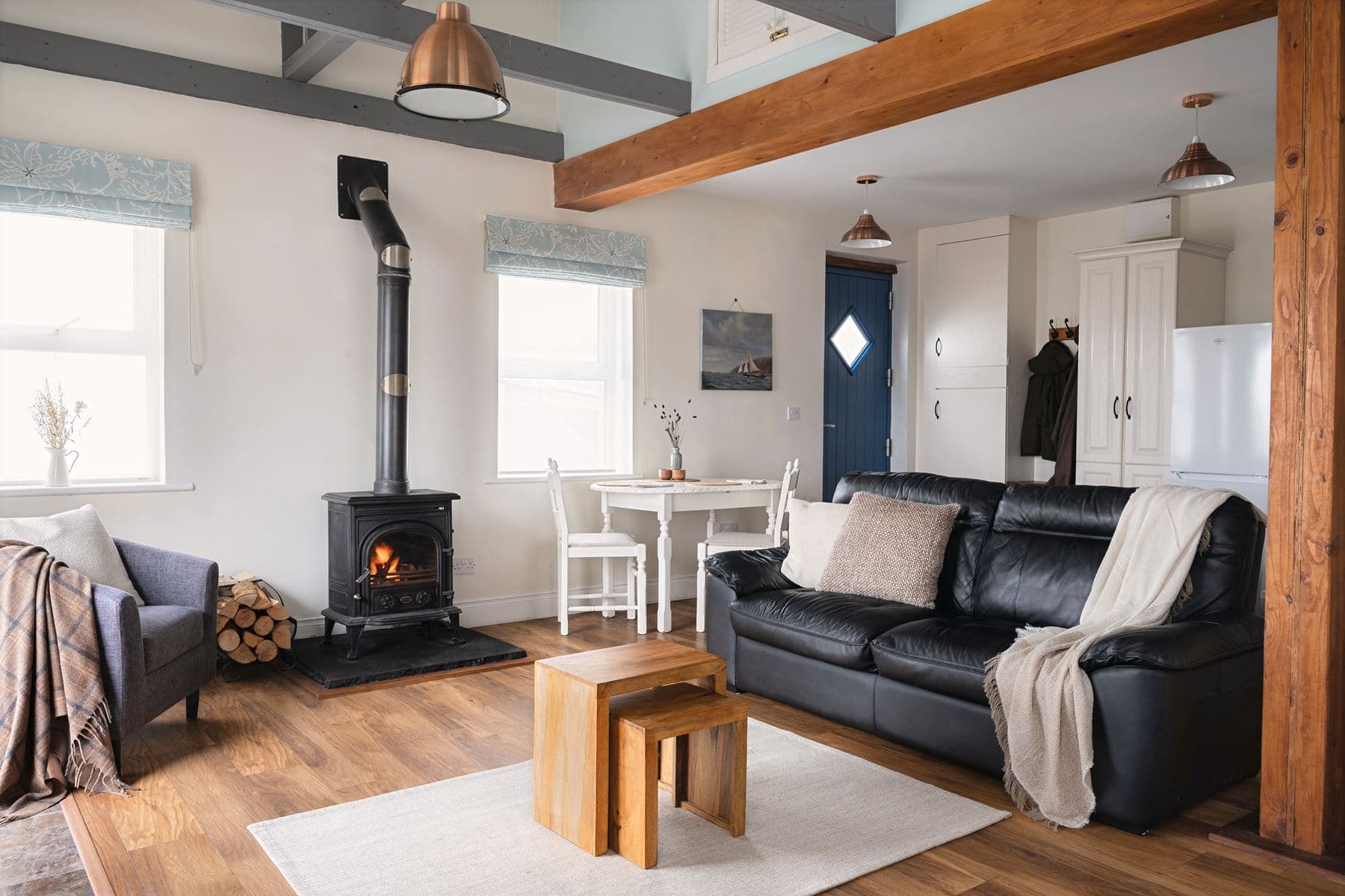 Spacious living room at The Studio featuring a log-burning stove, round dining table, and black leather sofa, with views toward the kitchen and entrance.