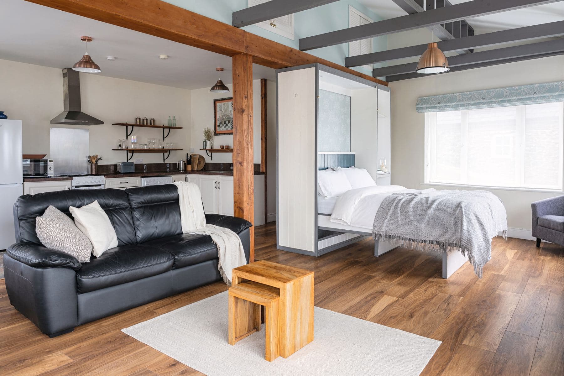 Open-plan studio layout with visible kitchen, black leather sofa, and a double bed positioned under a large window with natural light.