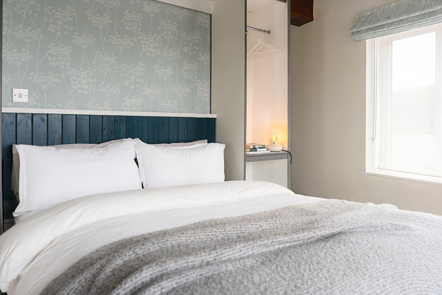 Calm bedroom space in The Studio with a blue wood-panelled headboard, soft floral wallpaper, and built-in wardrobe beside a large window.