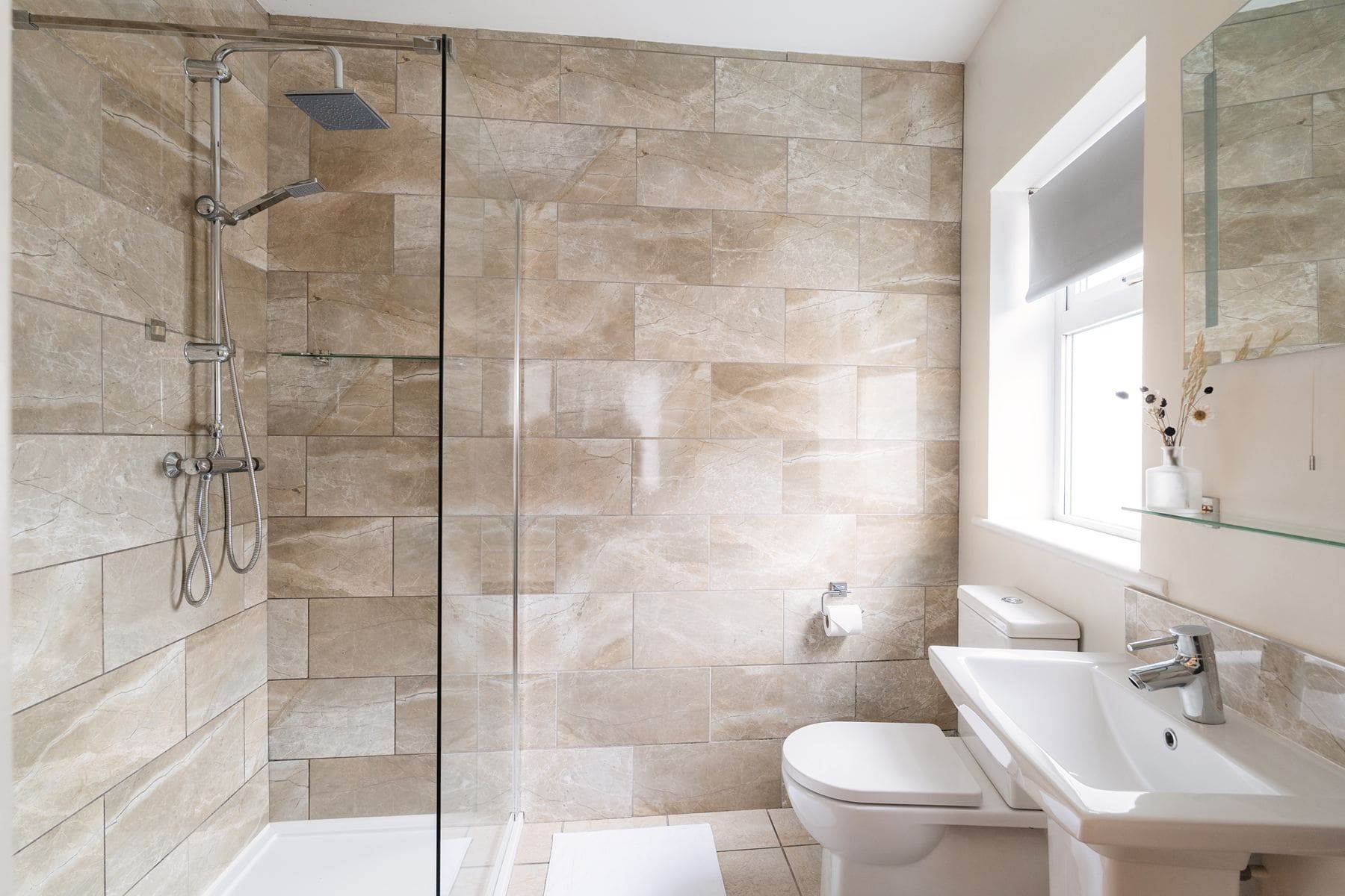 Modern bathroom with beige tiled walls, a walk-in glass shower, white toilet, and basin, naturally lit by a window.