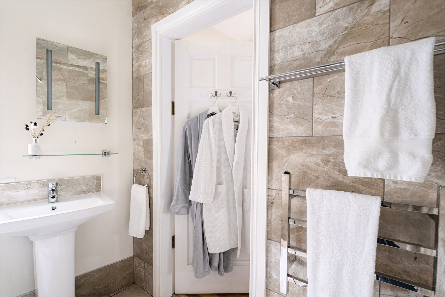 Bathroom interior featuring a towel rail, mirror, pedestal sink, and dressing gowns hanging on the back of the door.