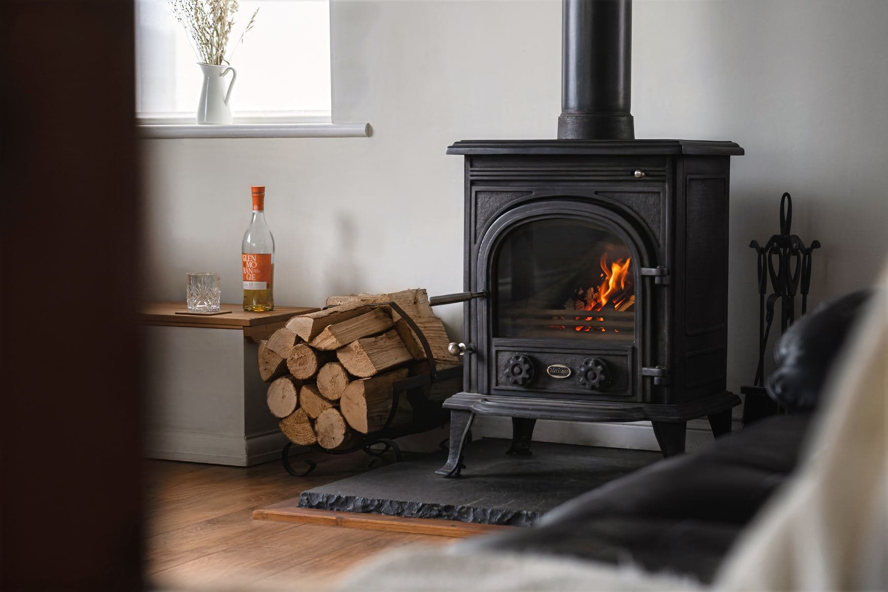 Close-up of a lit wood-burning stove with a stack of logs and a bottle of wine on a bench beneath the window.