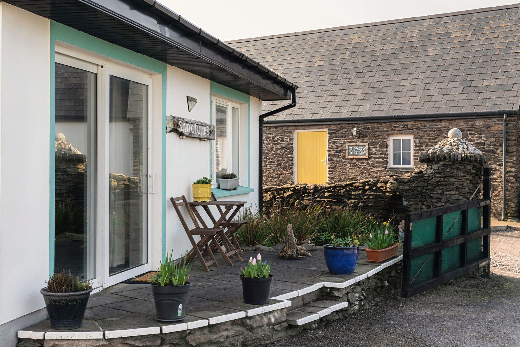 View of The Studio’s stone patio with outdoor seating and flower pots, facing a stone courtyard and the entrance to Pipers Cottage with its distinctive yellow door.