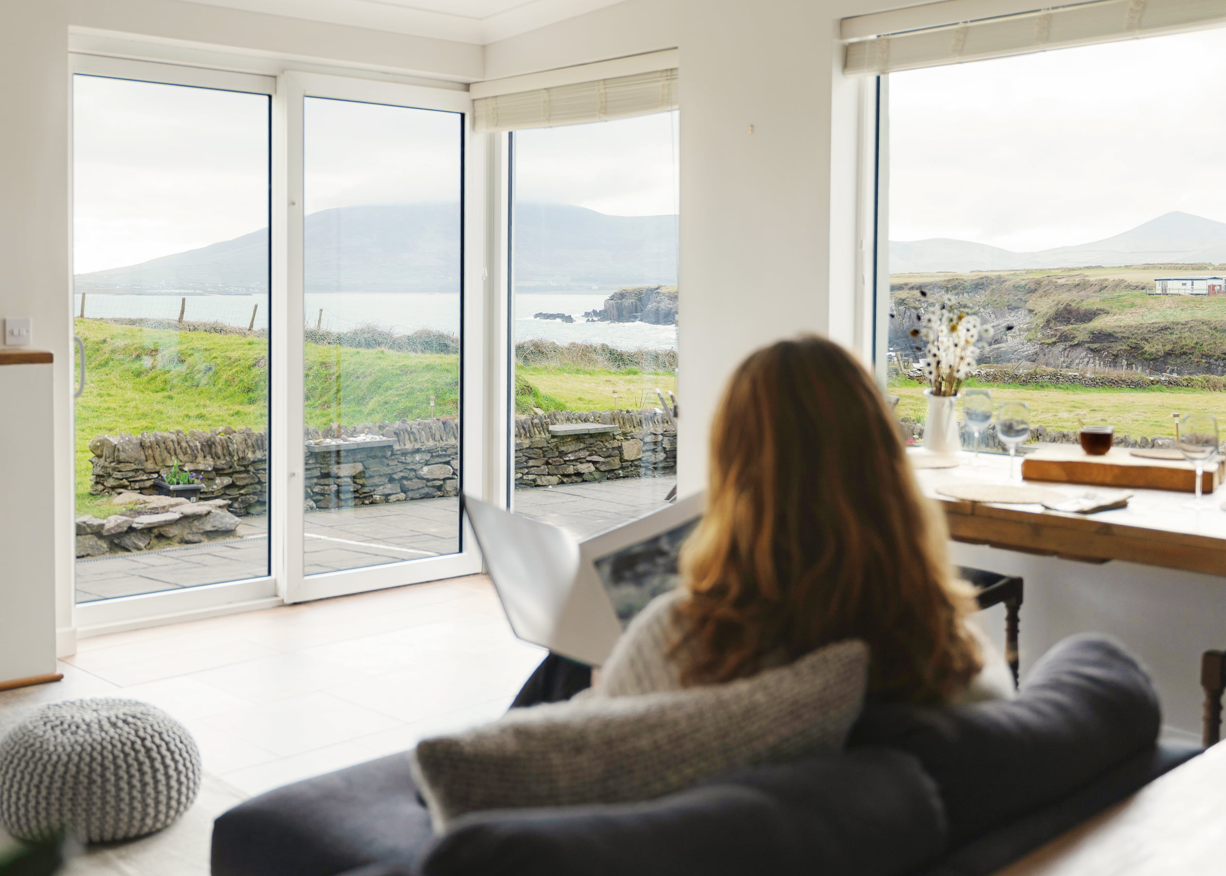 sanctuary-cottage-ocean-blue-panoramic-view-2880×1800 A guest sits on a sofa reading a book, facing large windows that look out onto grassy cliffs, stone walls, Ventry Bay, and Mount Eagle in the distance.
