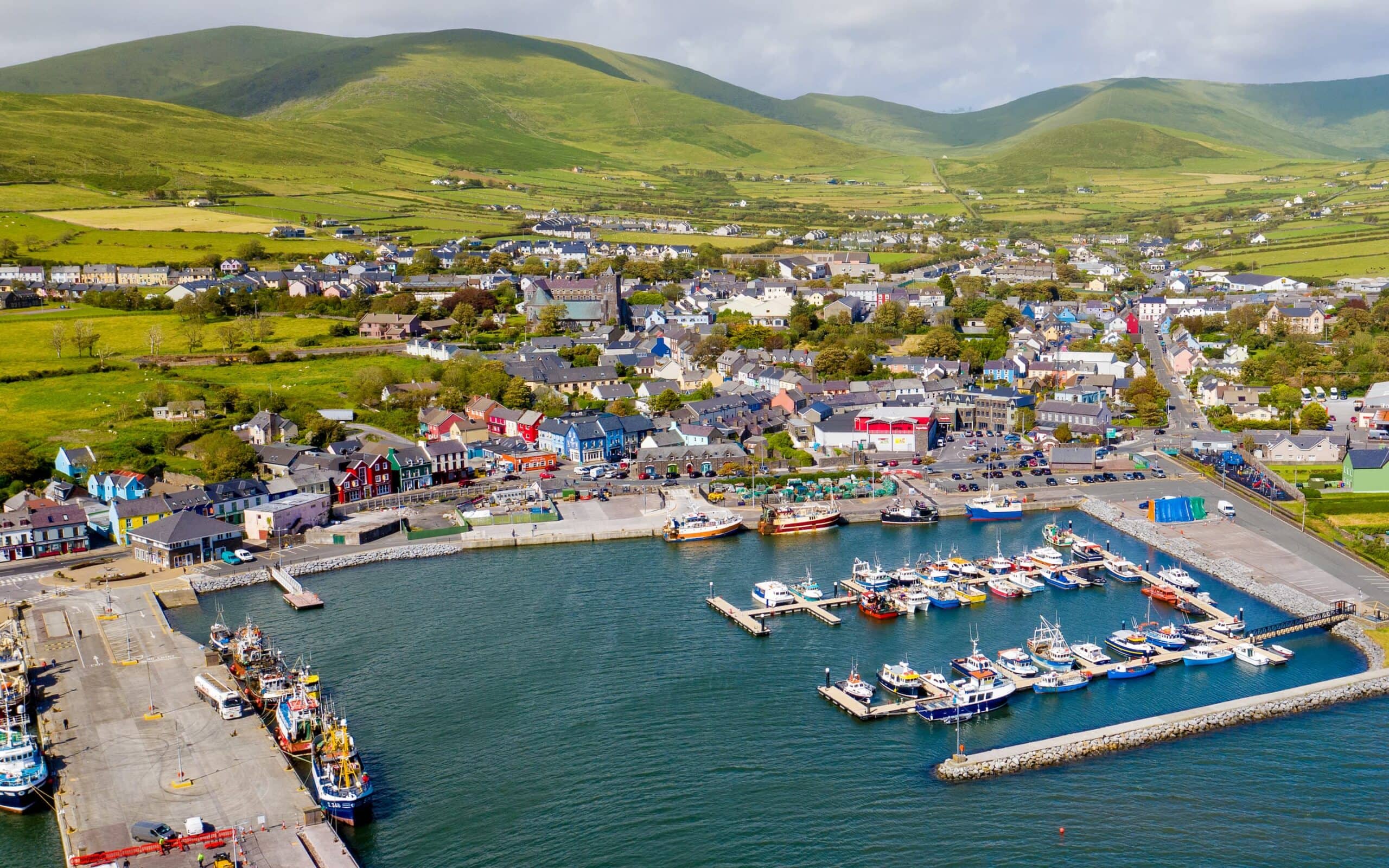 Aerial view of the vibrant village of Dingle in County Kerry, Ireland, showcasing colourful buildings, fishing boats, and rolling green hills in the background.