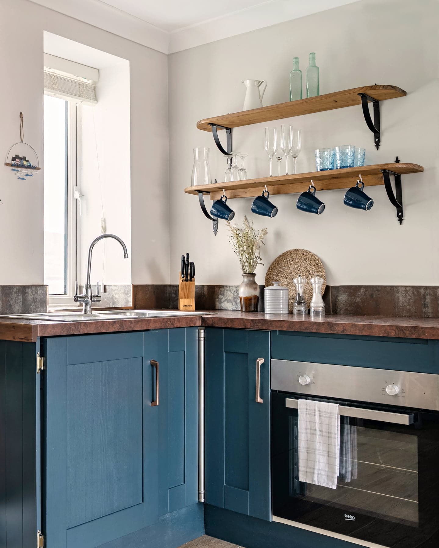 sanctuary-cottages-ocean-blue-kitchen-detail-1440×1800 Stylish, fully equipped kitchen at Ocean Blue, Sanctuary Cottages in Dingle, featuring deep blue shaker cabinets, open wooden shelving with glassware, and a modern electric oven.