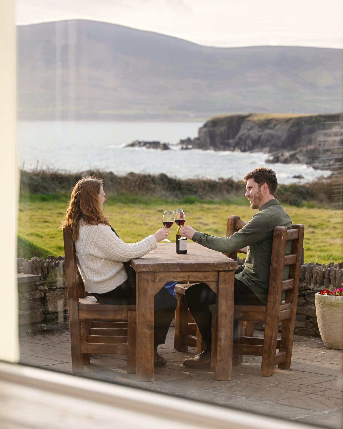 sanctuary-cottages-ocean-blue-window-patio-view-1440×1800 Couple enjoying wine on the private patio at Ocean Blue, Sanctuary Cottages in Dingle, with panoramic views across Ventry Bay and the dramatic cliffs of the Dingle Peninsula.