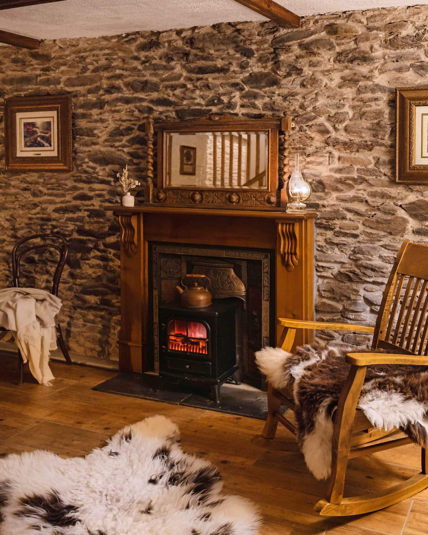 sanctuary-cottages-pipers-cottage-living-room-01 Warm and rustic living room at Pipers Cottage, featuring a traditional stone wall, wood-burning stove, antique mirror, and rocking chair with sheepskin throws.