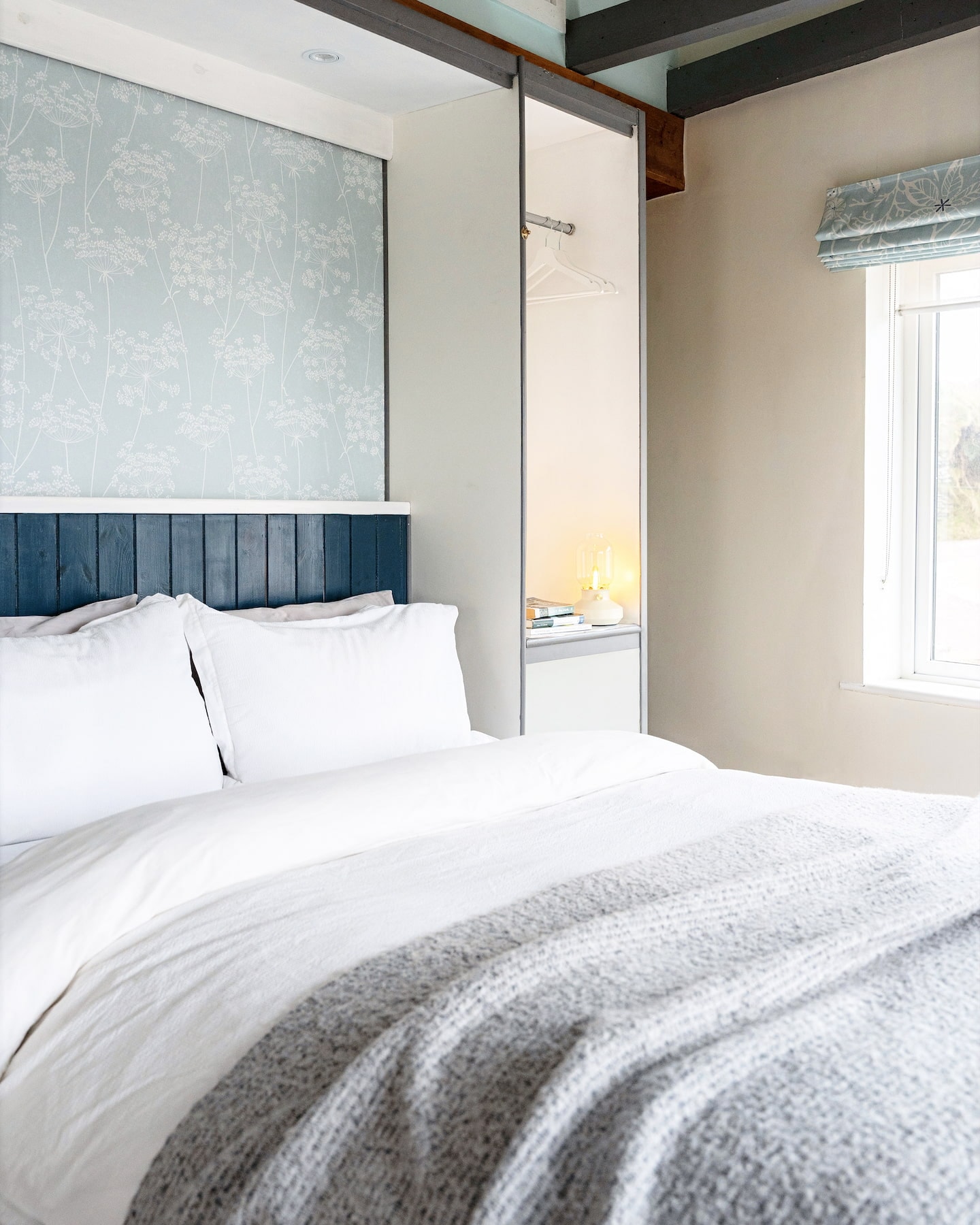 sanctuary-cottages-the-studio-bedroom-1440×1800 Bright bedroom in The Studio at Sanctuary Cottages, Dingle, featuring a double bed with white linens, blue wood-panelled headboard, floral wallpaper, and built-in wardrobe with open shelving and hanging space.