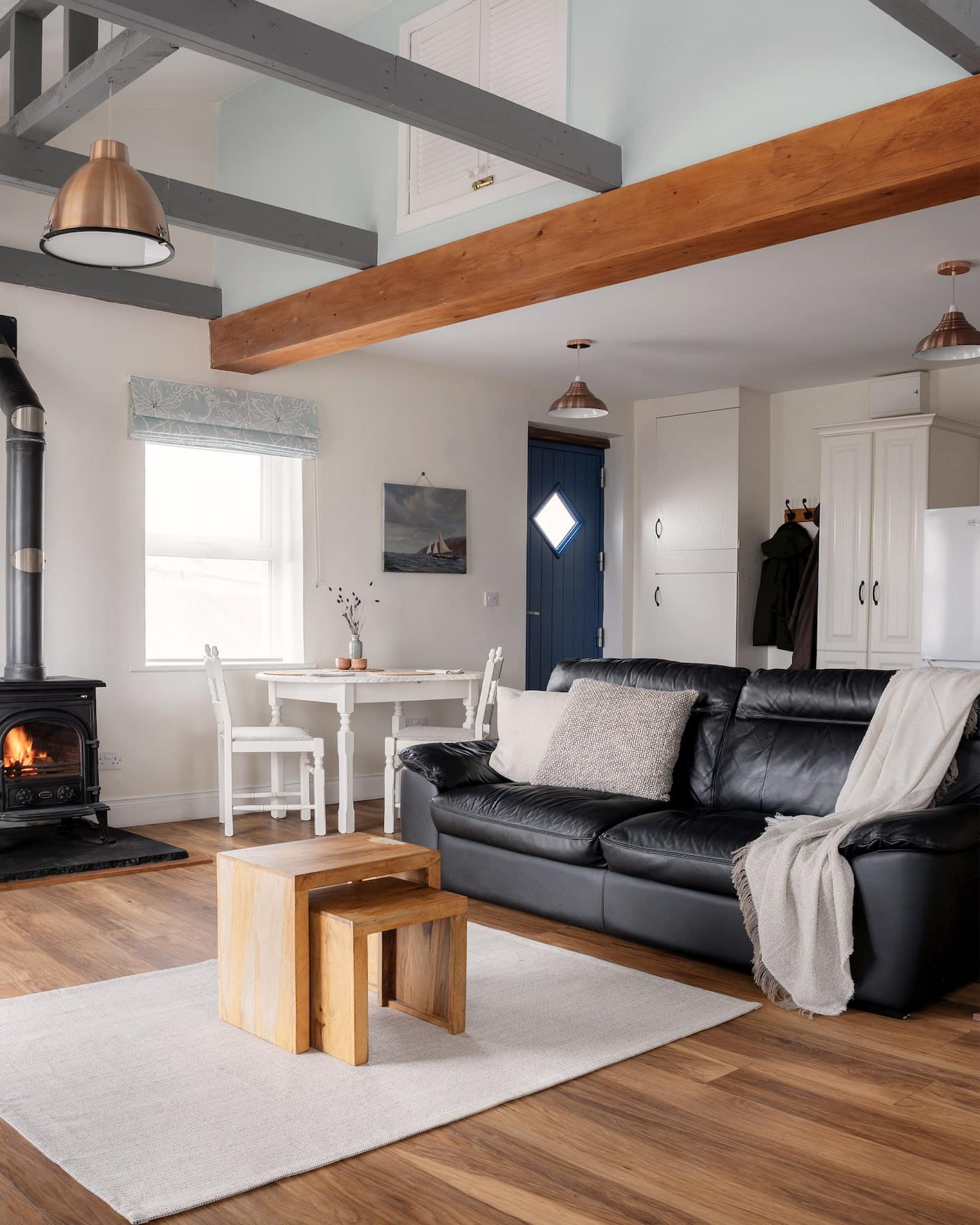 sanctuary-cottages-the-studio-living-room-angle-1440×1800 Spacious open-plan living area with a black leather sofa, wood-burning stove, dining nook, and natural light from multiple windows, accented by timber floors and vaulted ceiling beams.