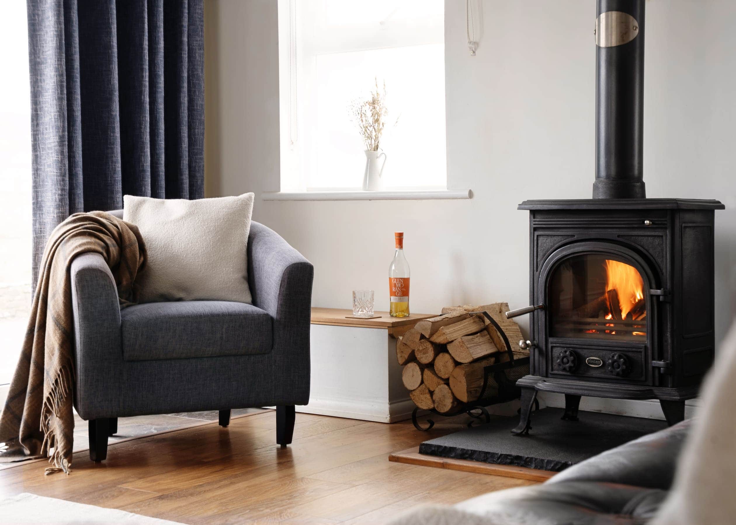 sanctuary-cottages-the-studio-living-room-hero-02 Cosy corner of The Studio at Sanctuary Cottages with a dark grey armchair, soft cushion, blanket, and a wood-burning stove set against a white wall with natural light from the window.