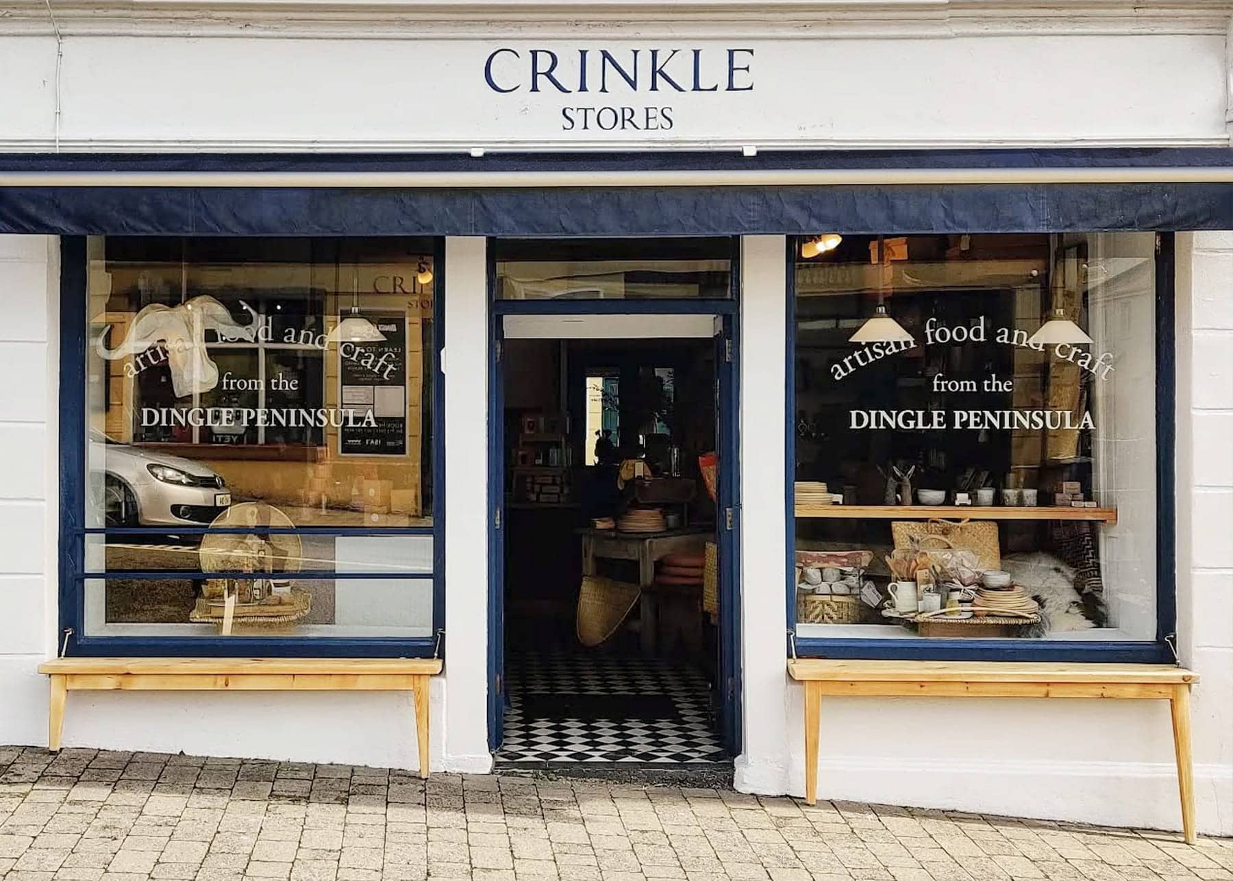 Storefront of Crinkle Stores in Dingle with white facade and navy blue trim. Two large display windows showcase artisan food and craft items from the Dingle Peninsula, with wooden display tables outside and black and white checkered floor visible through the open doorway.