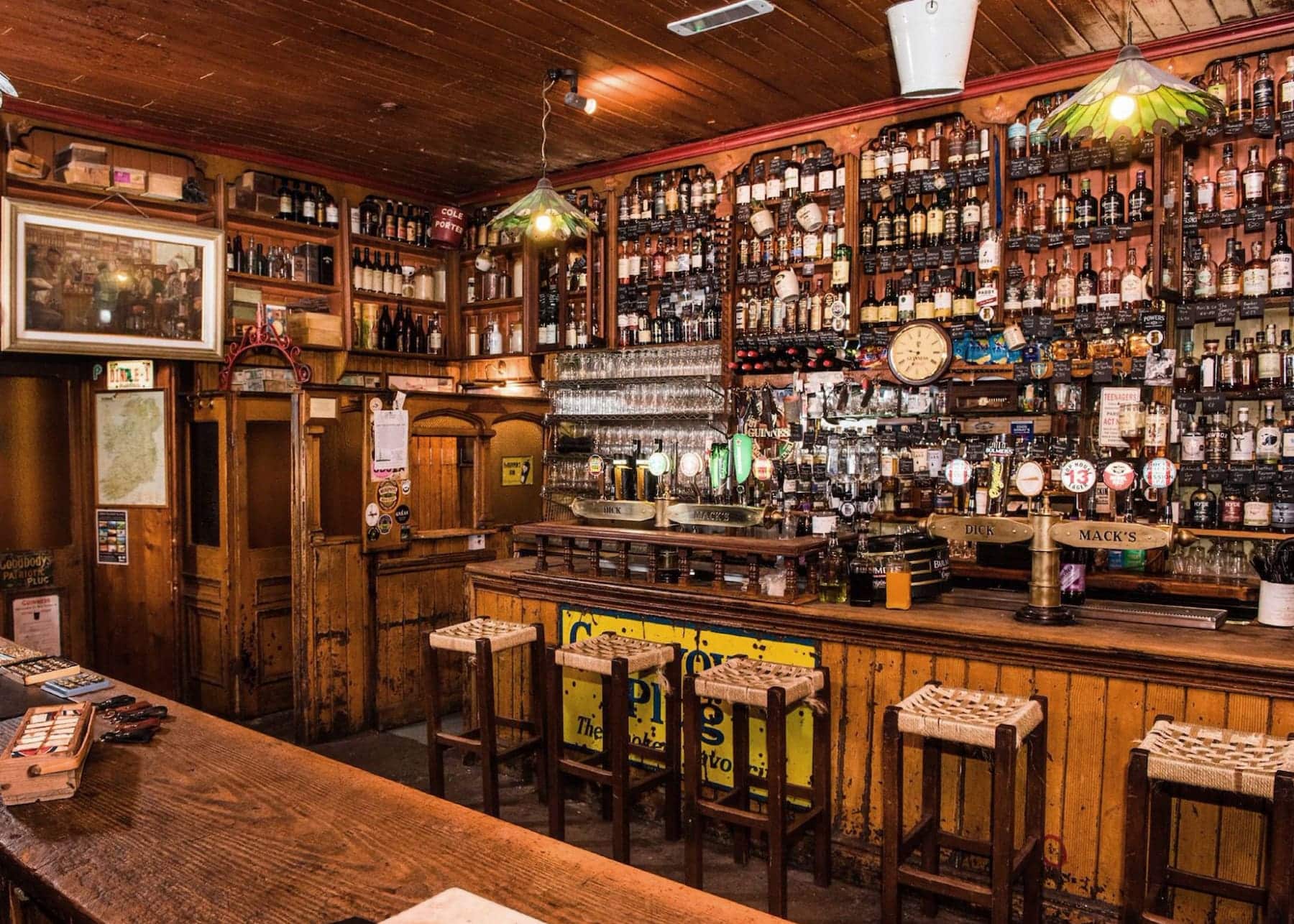 Interior of Dick Mack's traditional Irish pub in Dingle showing wooden bar counter with extensive whiskey collection on shelves, beer taps, vintage pendant lighting, and rustic wooden bar stools